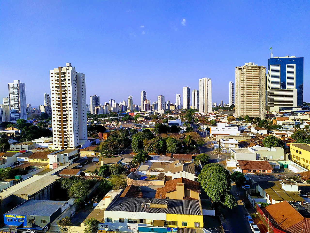 Vista de Goiânia, Goiás, com prédios altos ao fundo, casas e ruas arborizadas em primeiro plano sob céu azul